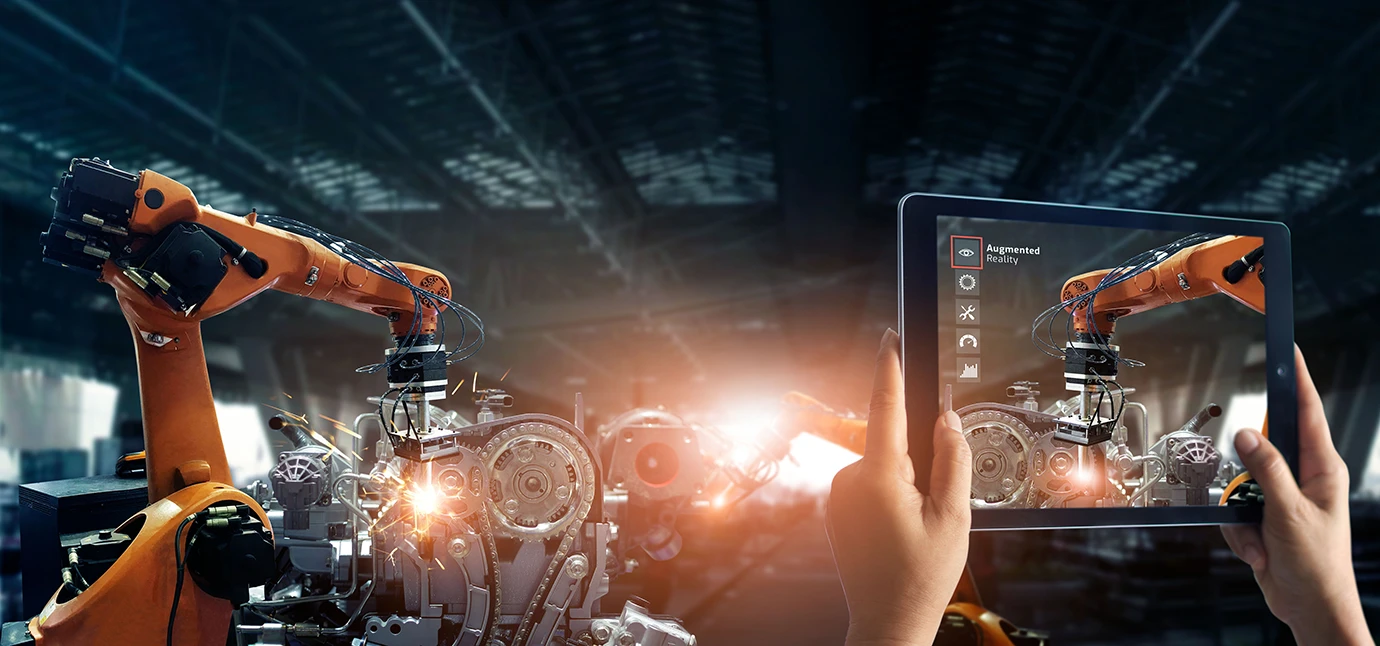 A person holds a tablet displaying augmented reality controls while observing robotic arms assembling machinery in a factory setting, with sparks flying from the equipment.