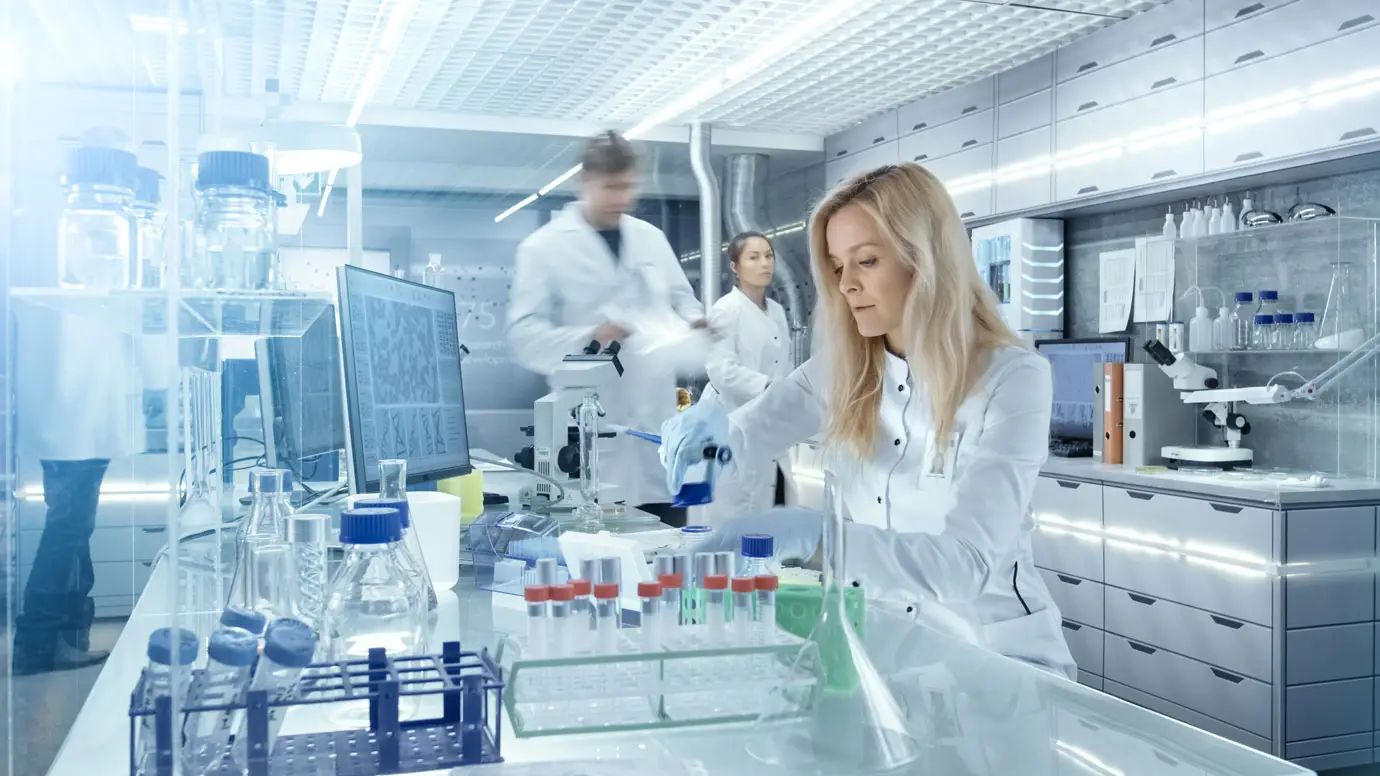 A woman in a white lab coat works at a lab bench with test tubes and scientific equipment, while two other scientists move in the background of a modern, high-tech laboratory.