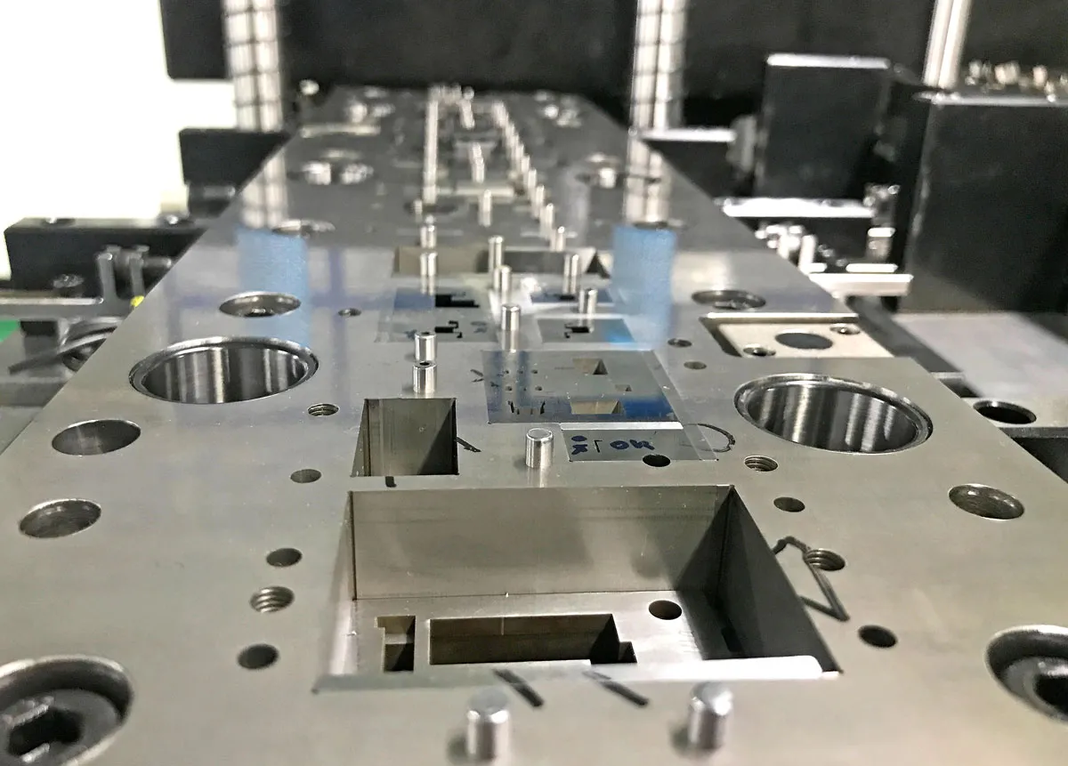 Close-up of a precision metal mold or die used in manufacturing, showing various holes, slots, and pins on a reflective metallic surface, with industrial equipment in the blurred background.