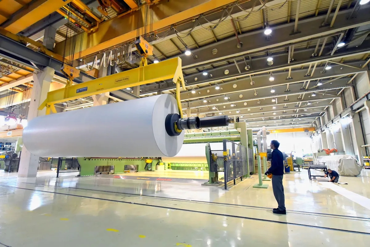 A large roll of paper is being lifted by an overhead crane in a bright, modern industrial facility. A worker in a helmet stands nearby, monitoring the process, while another person works in the background.