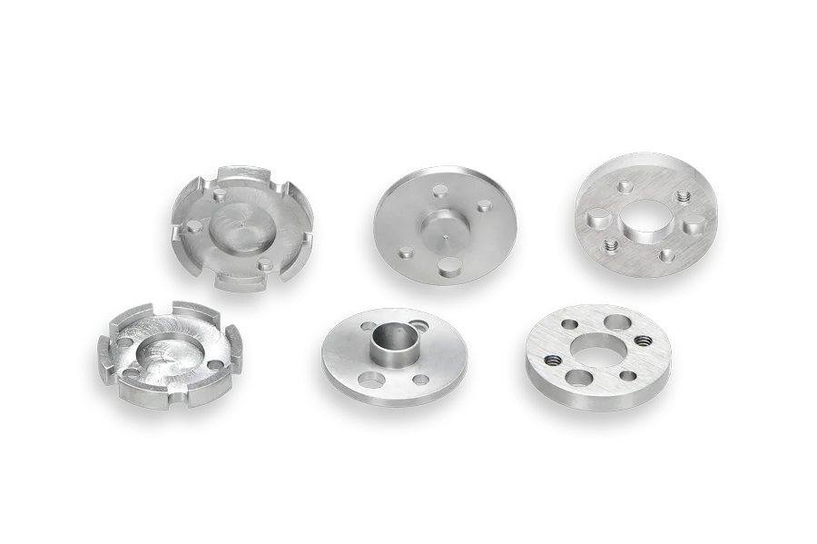 Six round, silver metal machine parts with various holes and cutouts are arranged in two rows on a white background. The parts have smooth surfaces and different internal and external features.